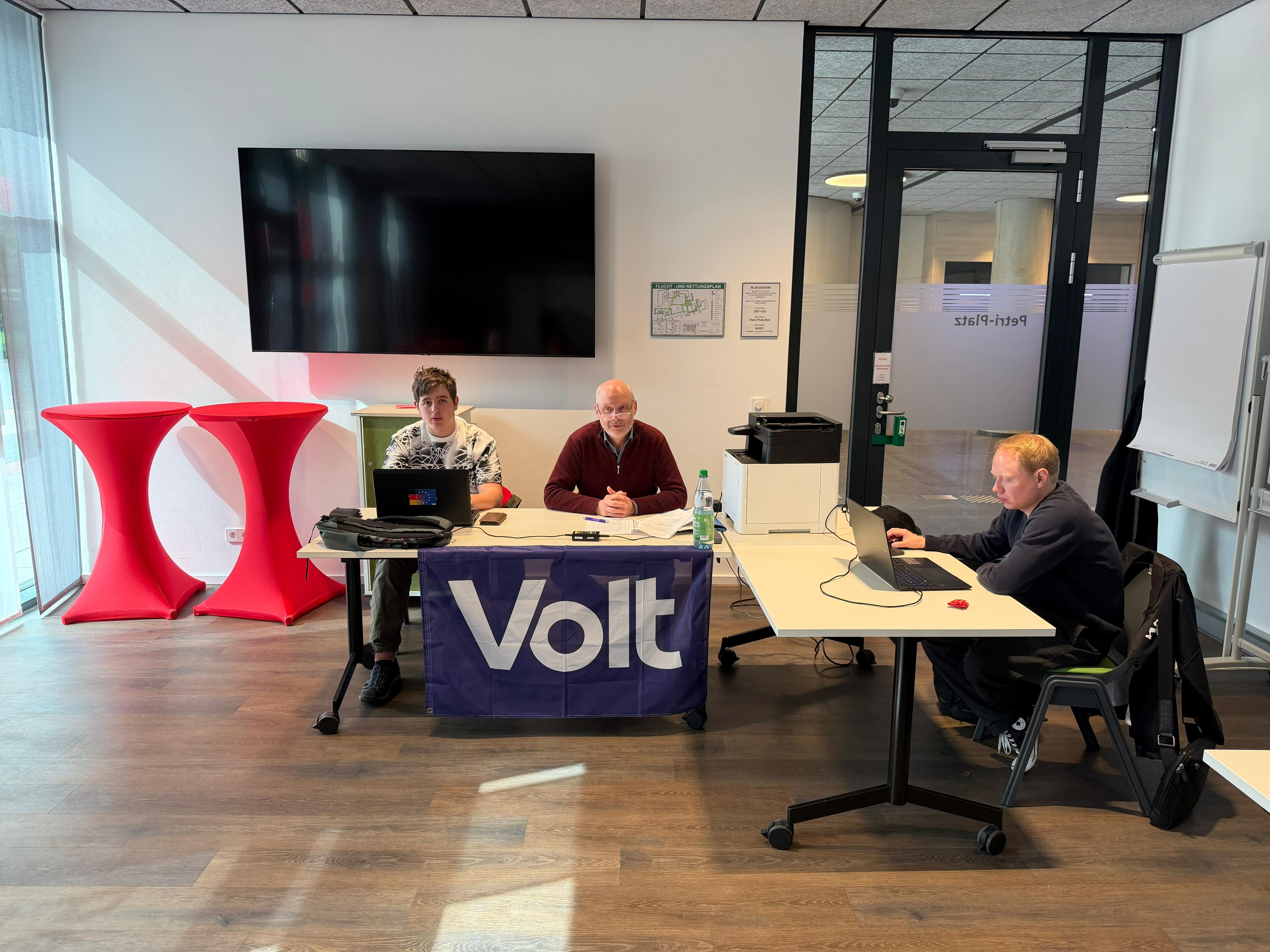 Drei Personen (Jack Ruder, Peter Gerken, Leonard Richter-Matthies) sitzen an Tischen mit Laptops und Unterlagen; vorne hängt eine Volt-Fahne.