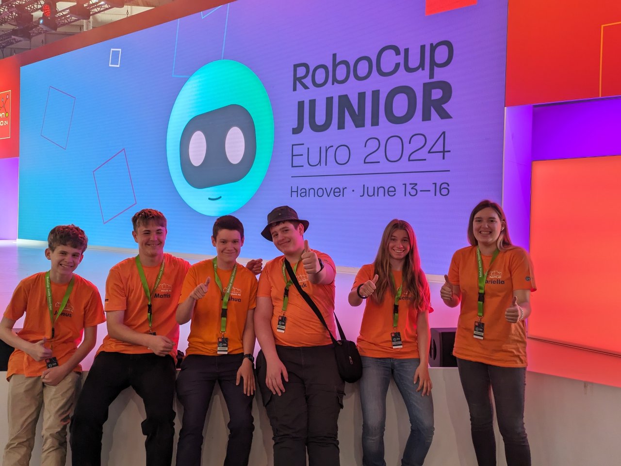 Sechs Personen in orangefarbenen Team-Shirts vor der Bühne des RoboCup Junior Euro 2024 in Hannover.