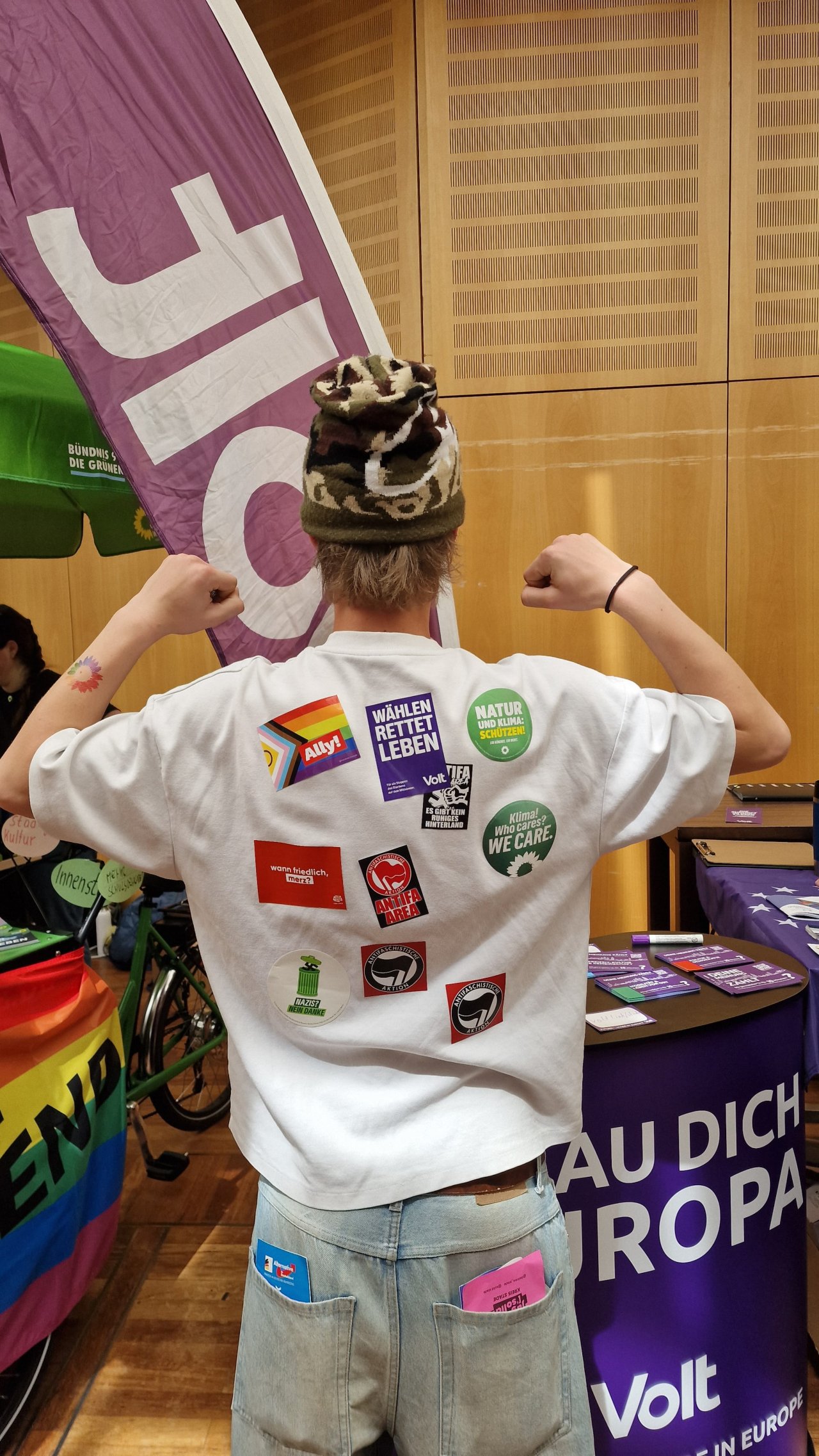 Person mit beklebtem weißem T-Shirt vor einem Volt-Banner, von hinten fotografiert.